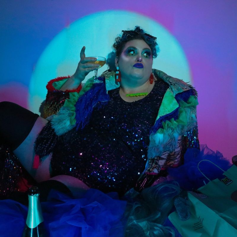 Deluxe Trashbag Cabaret - Aleksandra The Great, the shows MC, sits in a pile of trash with a glass of champagne in their hand.