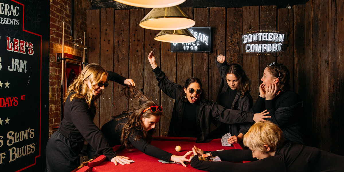 a group of 6 females pull and yell at each other as one of the females win the card game around the red pool table