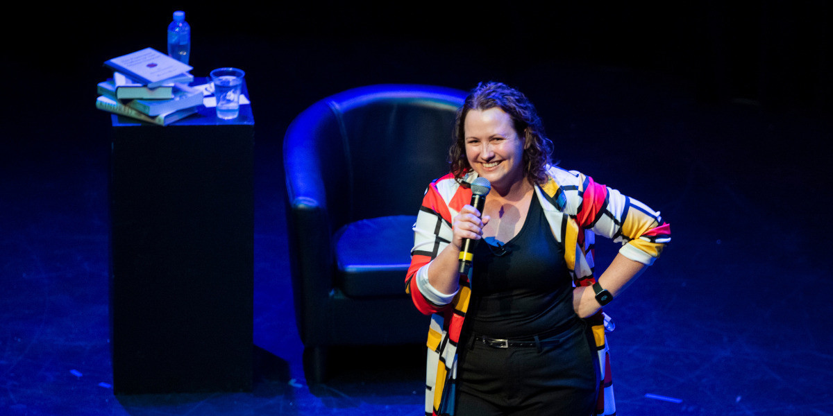 Archaeologist / comedian, KC Martin-Stone stands on stage, in a photo taken from a high angle of theatre seating. She has a beaming face as she listens to the audience laughter. KC is wearing black pants and top, with a striking Mondrian-style long light jacket over the top. The jacket is white with black lines creating square and rectangular spaces. Some of these spaces are randomly filled with red, yellow and blue blocks of colour. The stage lighting is blue, and blurred in the background are a chair and table with a pile of books and a glass of water.