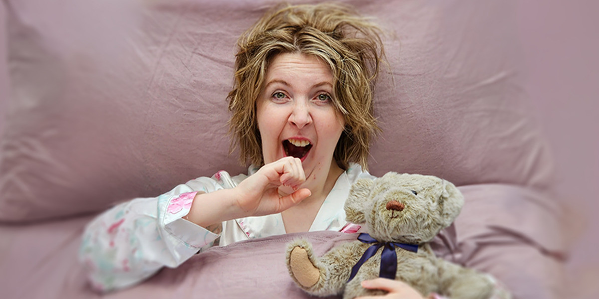 Snooze VR - A Chronic Fatigue Cabaret - A blonde white cis-woman comically yawning in bed, holding a teddy bear toy.