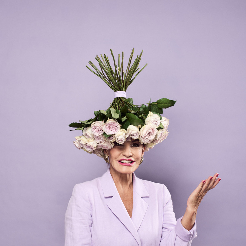 Comedian Tania Lacy sits facing camera, caught with a candid expression. She is wearing a lavender coloured suit, with a lavender coloured background and has a bunch of lavender coloured roses upside down on her head.