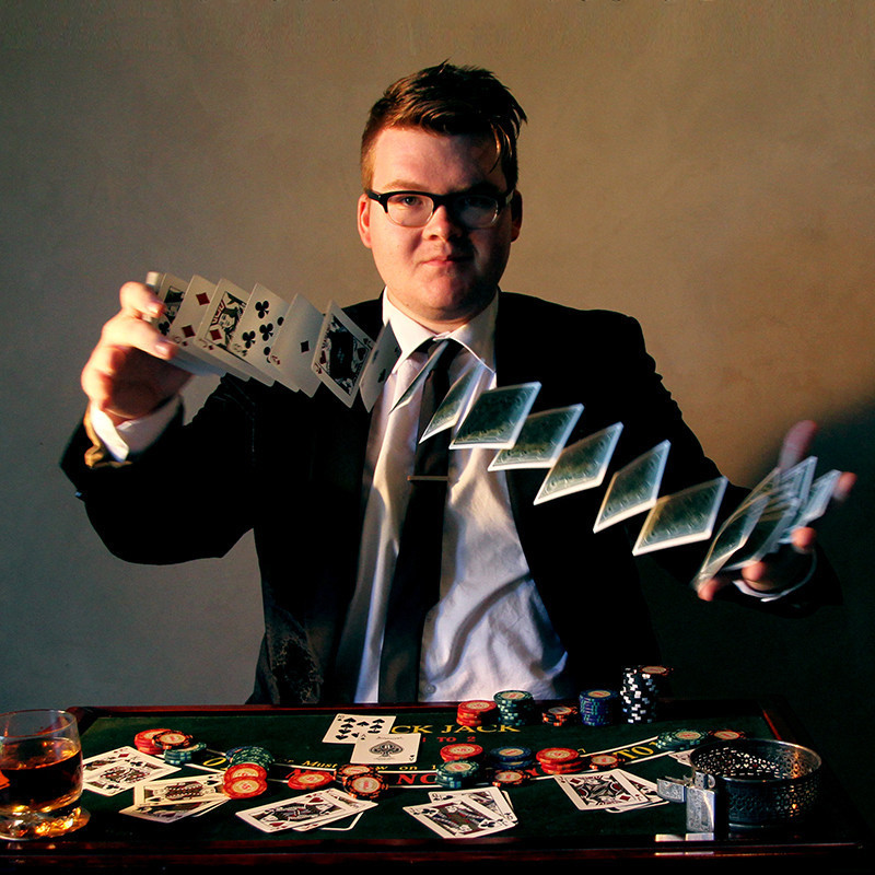 Josh Staley: The Conjurer at the Card Table - A photo of a man sitting down while shuffling a deck of playing cards with a serious expression on his face. In front of him is a table covered with playing cards and poker chips.