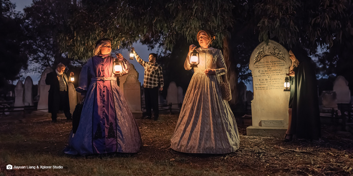 Two women dressed in late 1800's frocks, hold lanterns while standing in the foreground of grave stones in a Cemetery at dusk. The scene is lit only by the illuminance of the lanterns with figures outlined in the background.