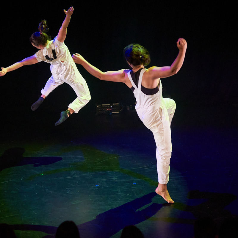 Two acrobats leap sideways together, horizontal to the ground with their backs toward the camera.