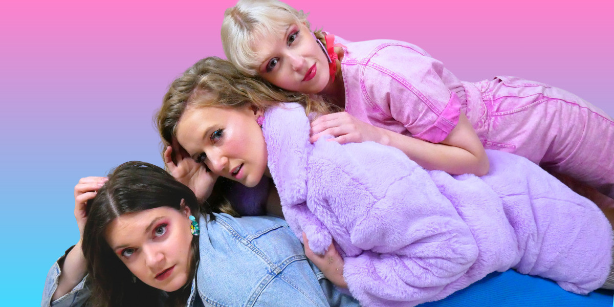 Three white women lay stacked on top of one another. The woman on the bottom has dark hair and is wearing a blue denim jacket. The woman in the middle has dark blonde hair and is wearing a fluffy purple coat. The woman at the top is wearing a pink boiler-suit and her platinum blonde hair is in plaits. They are staring into the camera with steely looks. Behind them is a gradient of blue, purple and pink.
