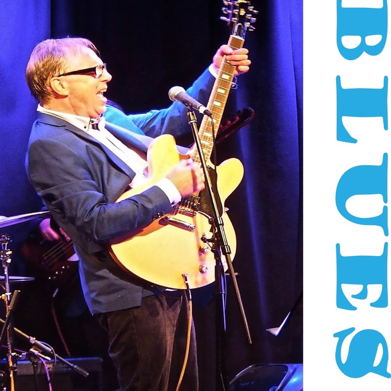 A photo of a man playing guitar on stage. He is wearing black framed glasses, a blue suit jacket, white shirt and black pants. There is text on the side of the image that reads ‘Blues’ in a light blue decorative font.