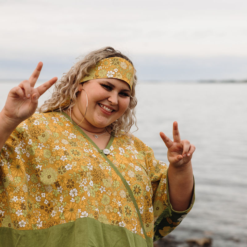 Young Fringe Artist dressed in flower power hippie clothing holding up double peace signs with a cheeky smile down at the beach.