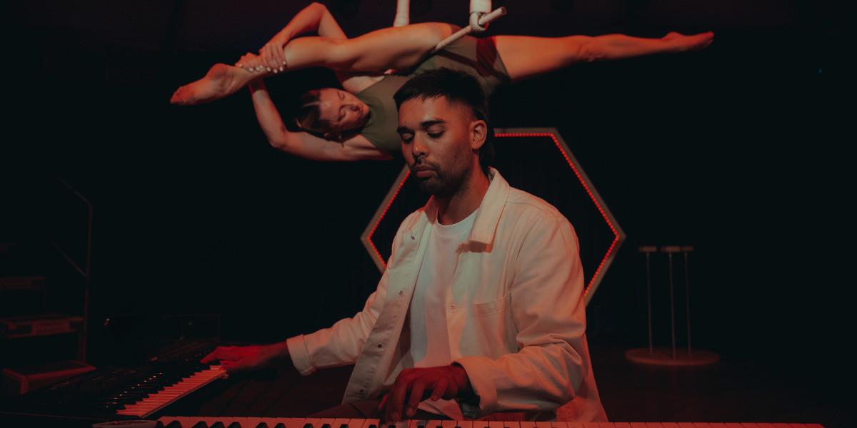 An acrobat performing the splits on a spinning trapeze high in the air, floating above a pianist who is sitting at two keyboards.