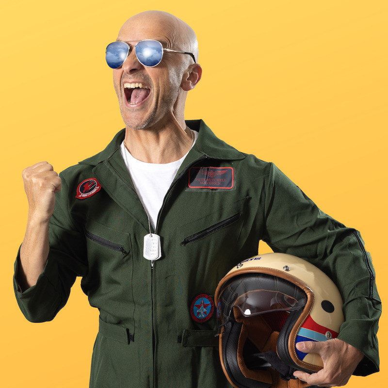 The performer with a bald head, but dressed like Tom Cruise in Top Gun, in a fighter pilot uniform, holding a helmet under one arm, and pumping his other fist, with his mouth open, yelling in excitement.