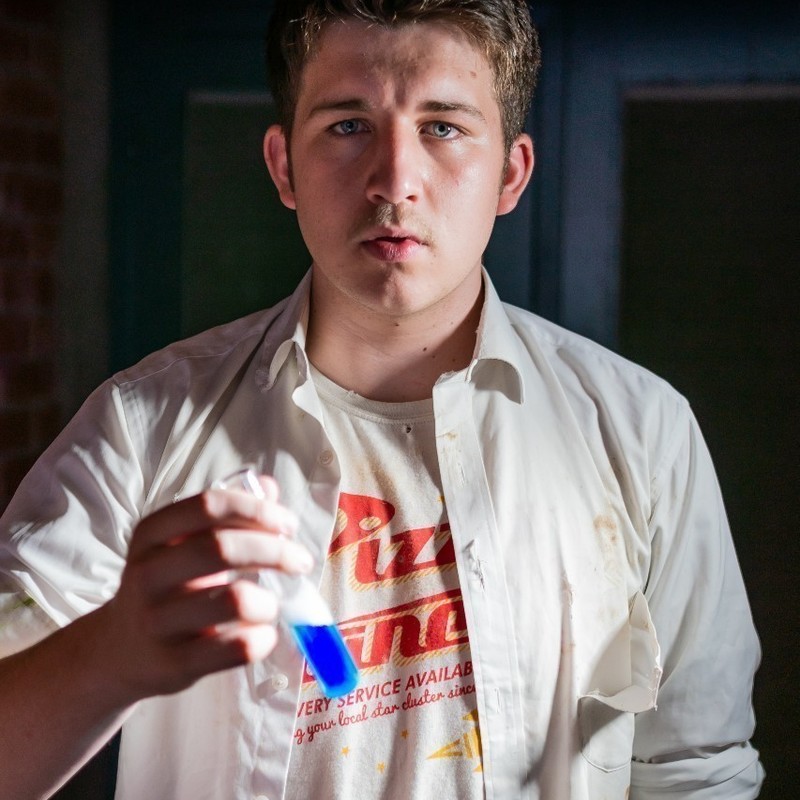 A photo of a young man with a serious expression on his face holding a test tube with blue liquid inside it. He is wearing a white t-shirt and white long sleeve shirt over the top.
