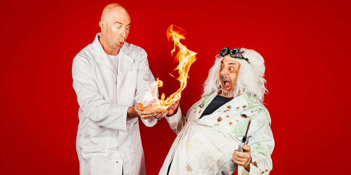Scientist Sam is very shocked by a big orange flame coming from his hands. Captain Chaos finds this hilarious as he leans back laughing holding a lighter.
Scientist Sam is dressed in a clean white science lab coat and Captain Chaos is dressed in a distressed white lab coat which appears to be burnt and covered in dirt, paint, slime etc. He also has a white Einsteinesk wig! The image sits on a vibrant red background.