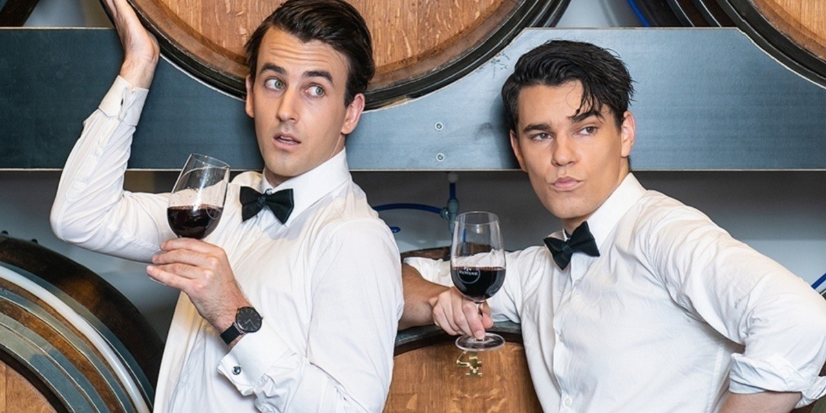 Two men in formal black tie stand in front of wine barrels looking curious and a little bit silly