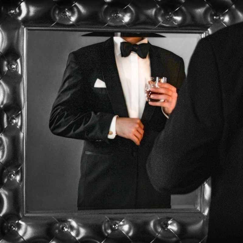 The Rad Pack - An image of a reflection of a person in a mirror wearing a black suit jacket, white shirt and black bow tie. They are holding a clear glass filled with brown liquid.
