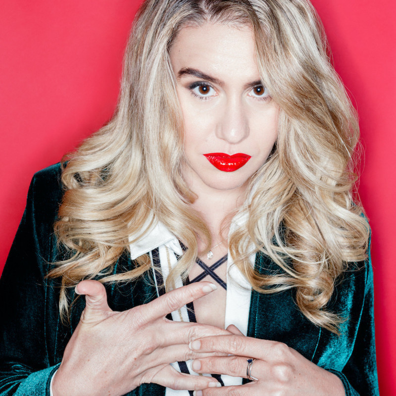 A performer with curled blonde hair and bright red lips stares intently at the camera with her chin down slightly and a neutral expression. She is wearing a dark green velvet jacket with a white shirt and black trim infront of a hot pink backdrop.