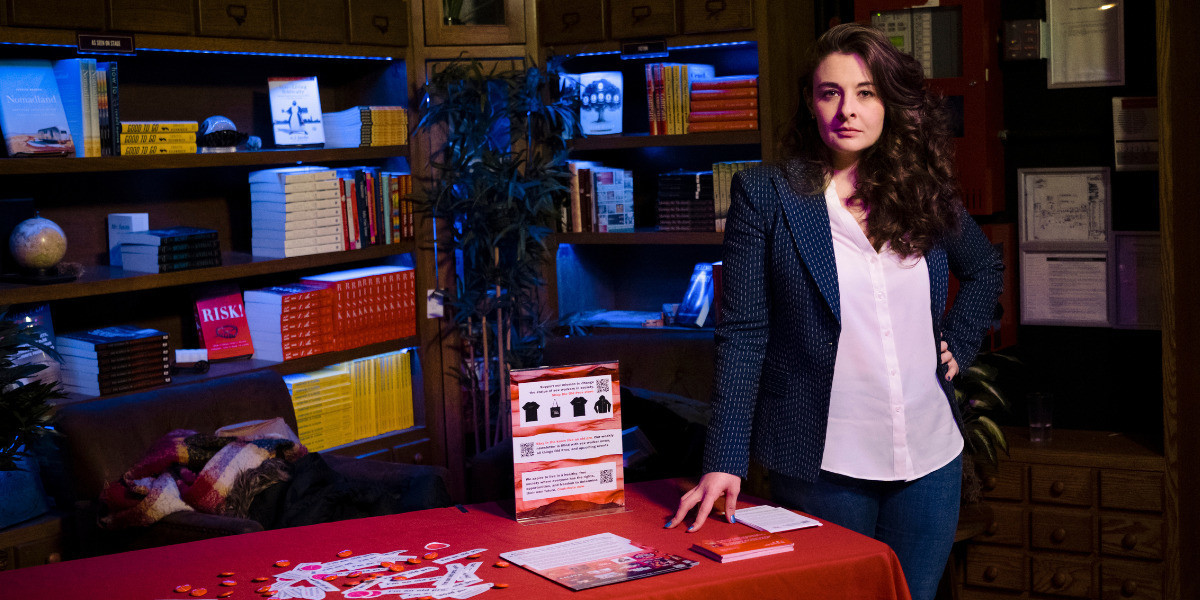 Kaytlin Bailey, mid-30s white cis-woman with brown long hair wearing a grey suit, stands next to a red table with bookshelves behind her