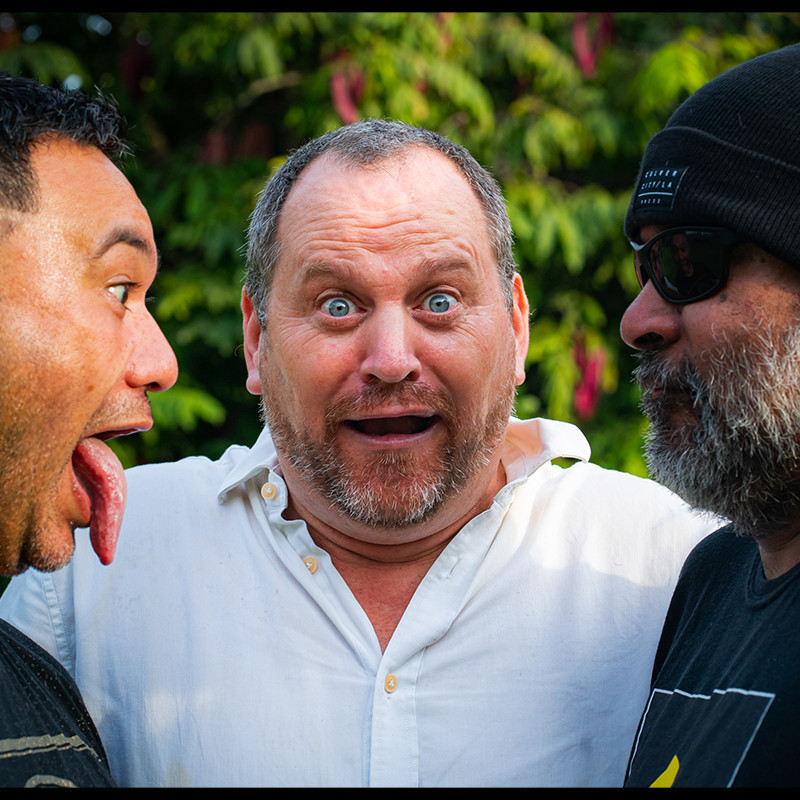A headshot photo of three men facing each other. The man on the left is poking his tongue out. The man in the middle has blue eyes and is wearing a white shirt with an animated facial expression. The man on the right is wearing a black beanie and sunglasses and is staring at the guy poking his tongue out.