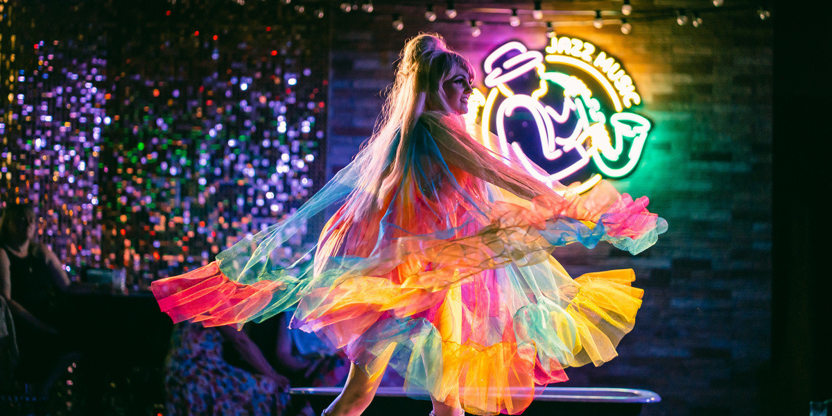 Blonde burlesque performer twirls on stage in colourful rainbow costume dress.