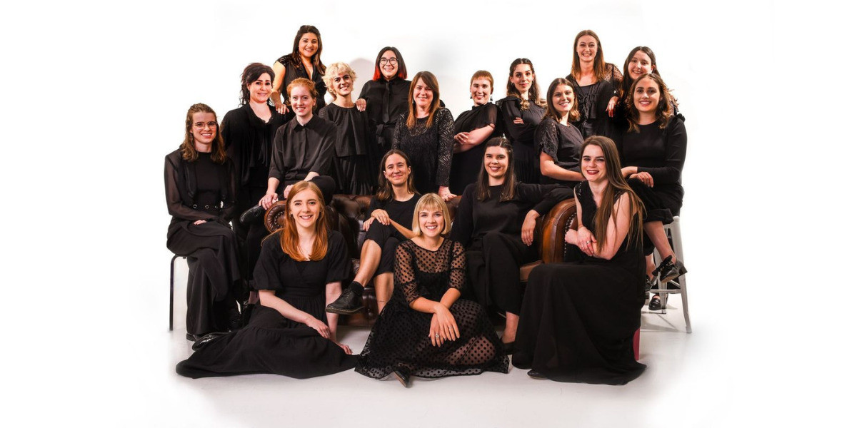 A group of women and non-binary people all wearing black are smiling and looking at the camera in front of a white background.