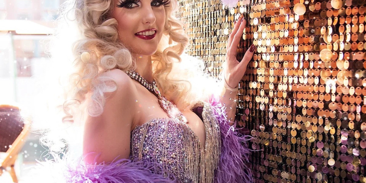 Blonde burlesque performer stands against gold glitter wall, wearing sparkly purple costume with a purple burlesque boa wrapped over arms.