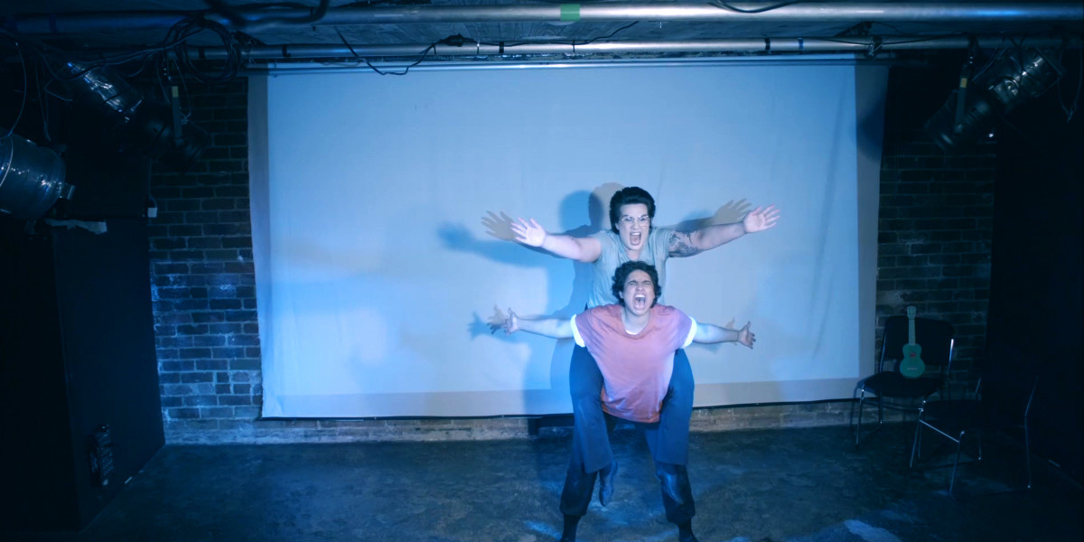 Dylan (Lou Sebial) on Solar (Neptune Henriksen)'s back, in a piggyback lift, with both holding hands out to the audience, and mouths open screaming.