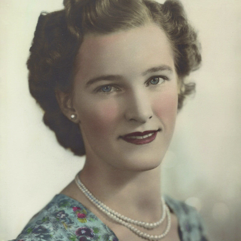 CANCELLED - Matt Byrne's: Mum's Story - A headshot photograph of a woman with short blonde hair and blue eyes. She is wearing pearl earrings, two pearl necklaces, and a blue floral dress. Her makeup is gentle and features pink blush upon her cheeks and dark red lipstick.