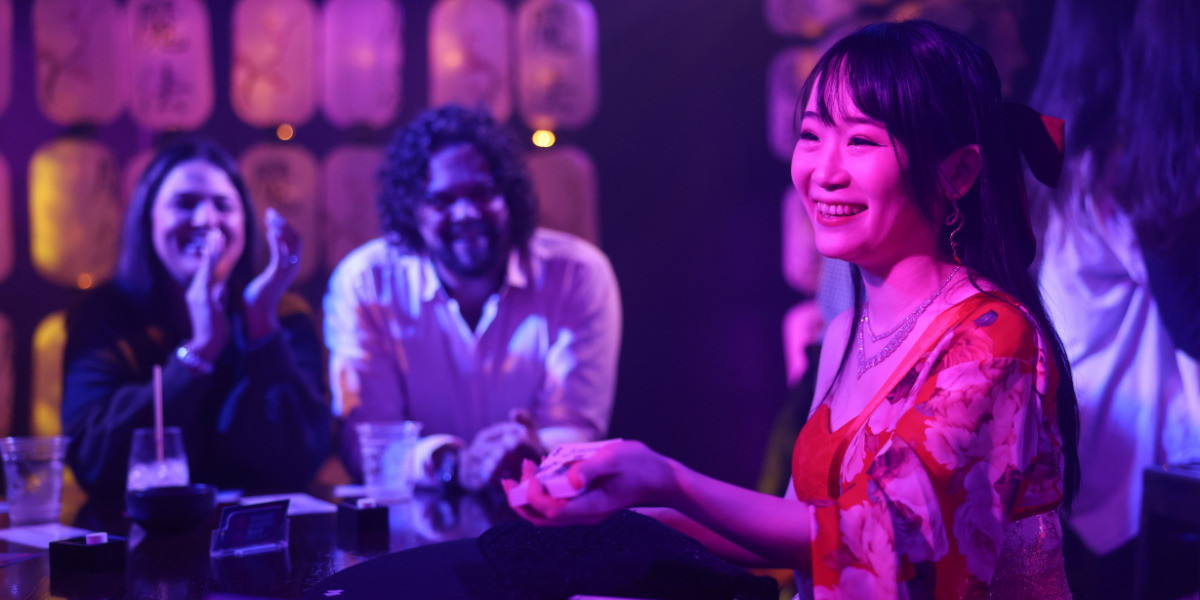 A Japanese magician holds out a pack of cards, smiling. Two people behind her are smiling and clapping