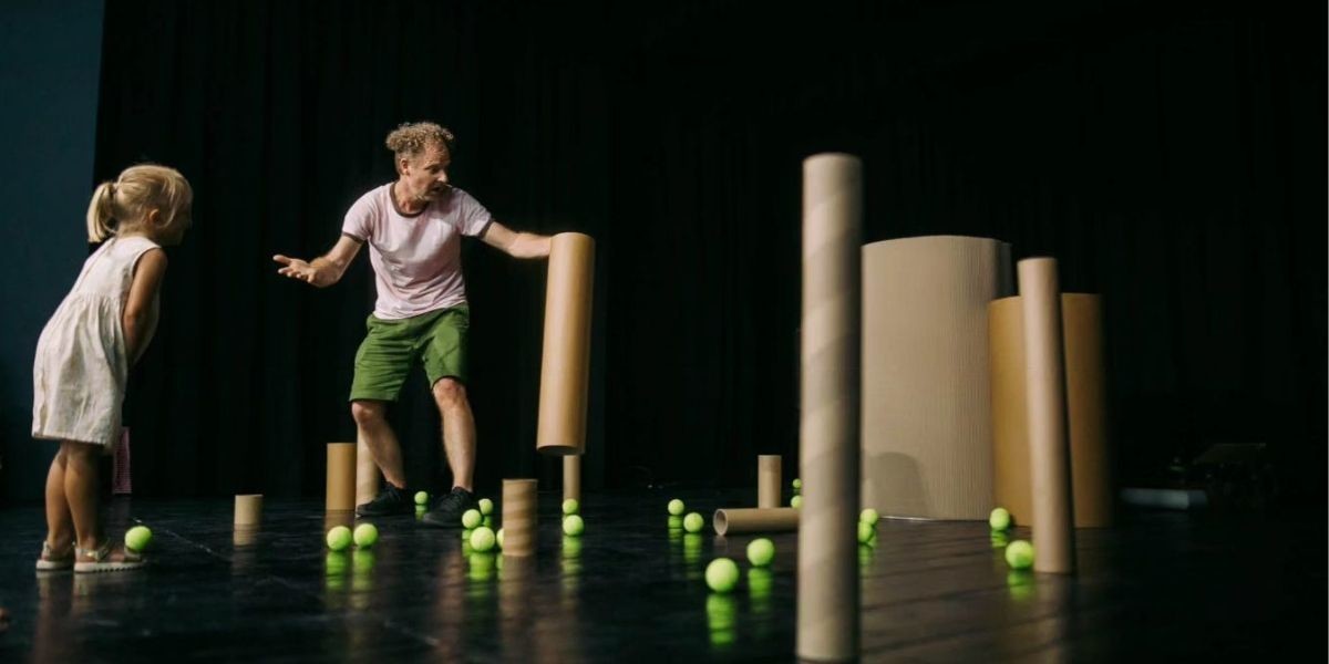 Horizontal brown cardboard tubes fill the space with many tennis balls scattered on the ground. A man and a 4 year old girl freeze in space looking at the balls.