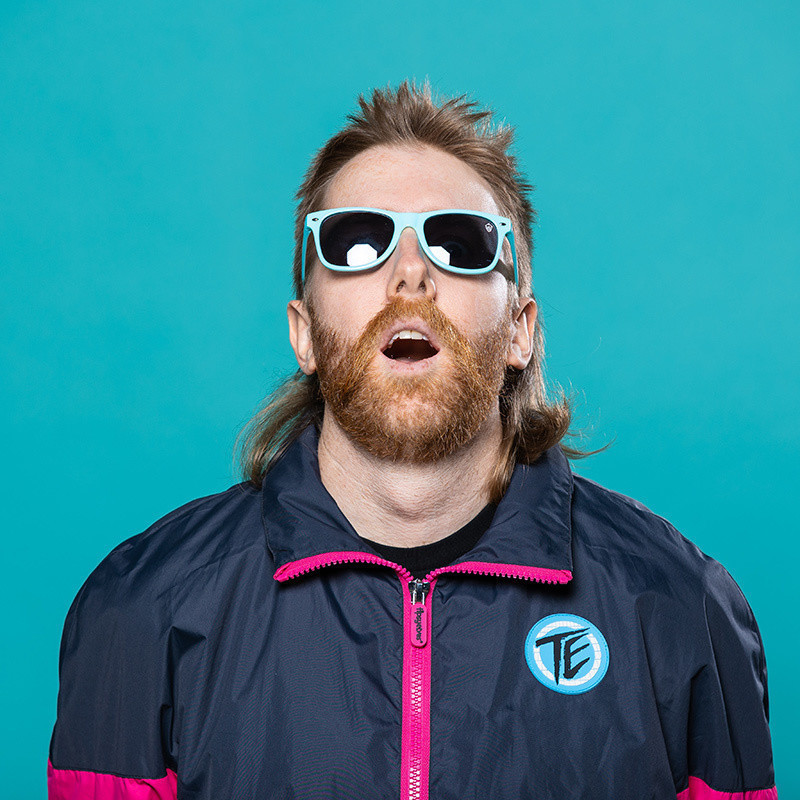 Matt Stewart comedian with red hair and mullet looking up in a pair of blue rimmed sunglasses with his bands behind his back and a pink and blue sports jacket