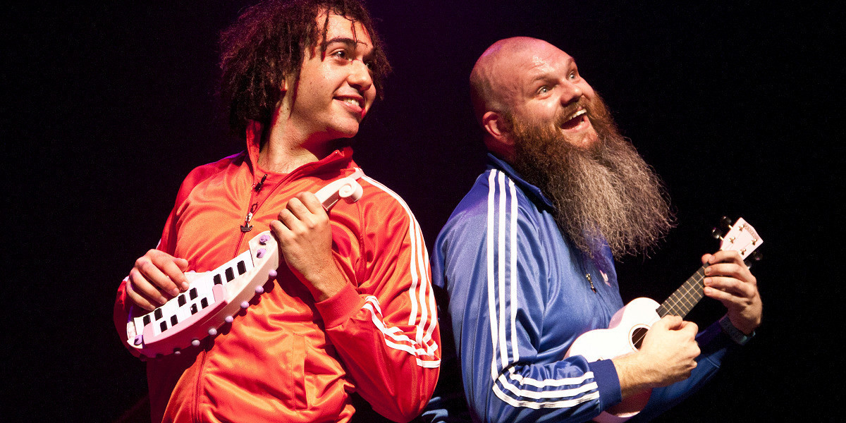 Gene Peterson is wearing a red tracksuit and holds a tiny pink keyboard. Adam Page is wearing a blue tracksuit and is playing a pink ukulele. Both artists look comically happy