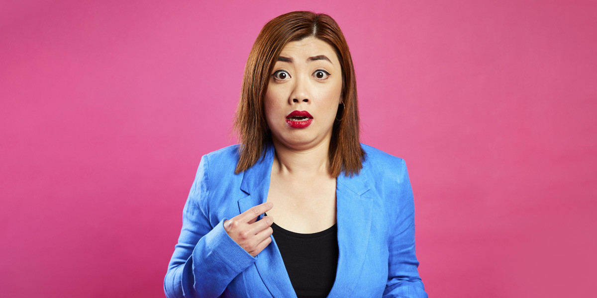 Ting Lim: Well This Is Awkward - Artist standing in front of a pink background holding onto the lapel of their jacket