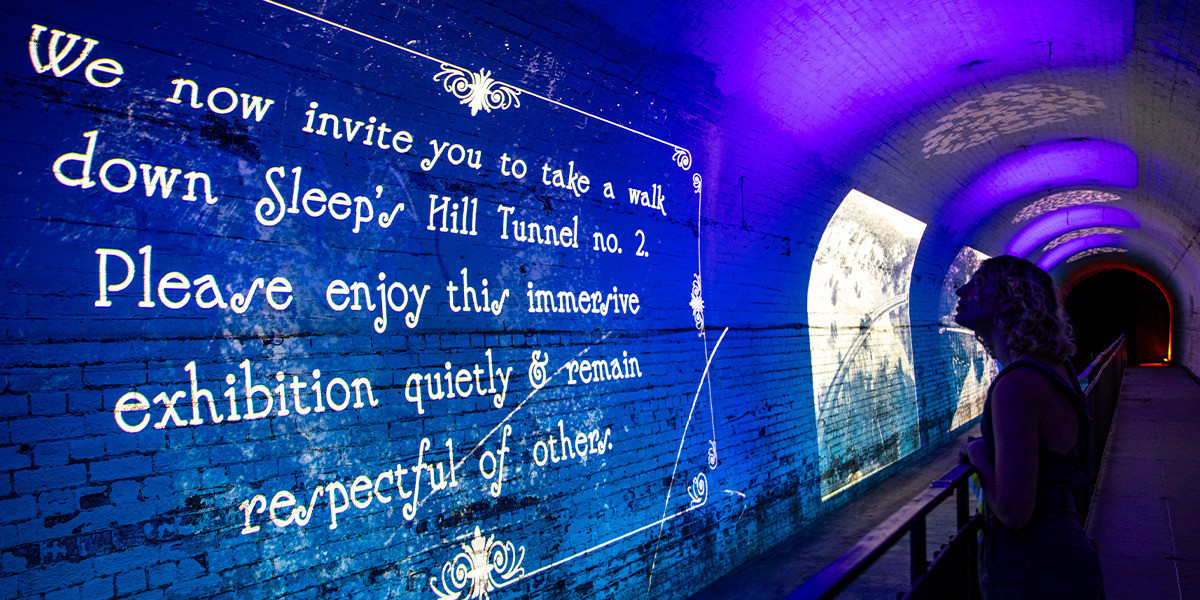 Sleep's Hill Tunnel - Brick tunnel wall with blue and purple lights projected onto it. White writing is on the wall reading: We now invite you to take a walk down Sleep's Hill Tunnel no. 2. Please enjoy the immersive exhibition quietly & remain respectful of others. A woman is leaning against the rail looking at the projection