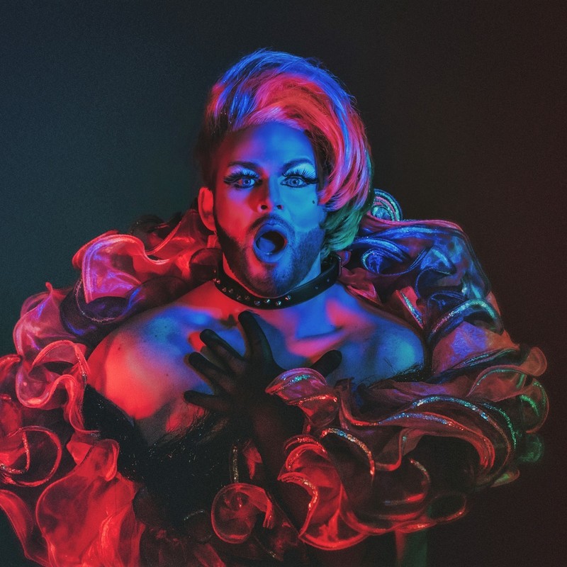 Drag performer Leather Lungs lit in neon red and blue, gasping in delight to see you