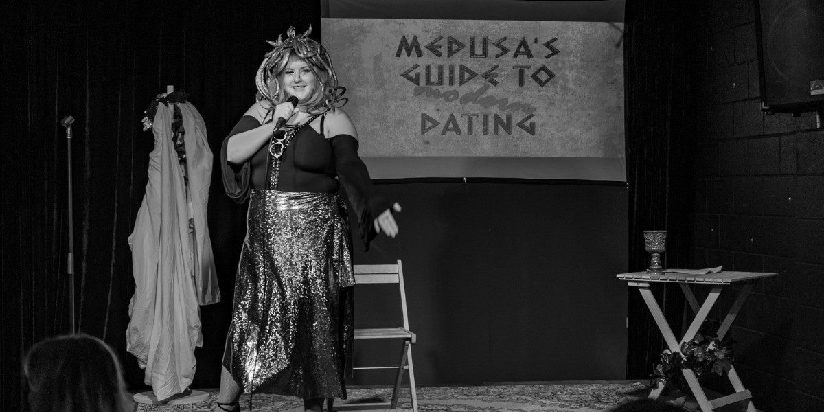 Medusa's Guide To Modern Dating - A black and white image of Medusa on stage. Medusa stands with her hair covered in snakes, dressed in a black lace-up top with flowing sleeves, and a reflective snake-skin pattern skirt, gesturing to the audience. She has heart-shaped glasses hanging from the top of her shirt. She has a smile on her face and is holding a microphone. Behind her is a screen that shows text saying 'Medusa's Guide to Modern Dating' in Ancient Greek style lettering, with the word 'Modern' laying on top in swirling lettering. To Medusa's left is a wooden coat stand with a white sheet hanging from it and a head wreath hanging from it. To her right is a small table covered in vines, with a goblet on the top.