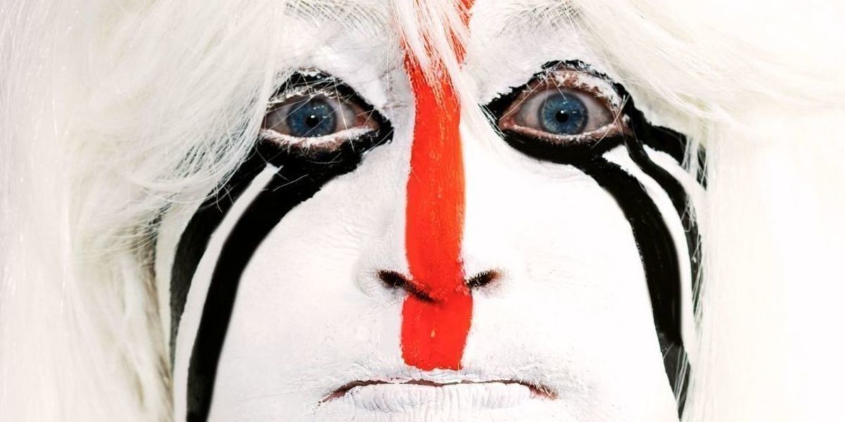 Kelly Mac stares at the camera wearing a long white wig and puffing her cheeks full of air. Her face is painted white with a thin red line from the centre of her forehead to her chin. Three wavy black lines run from the corner of her eyes to the edge of her cheeks.