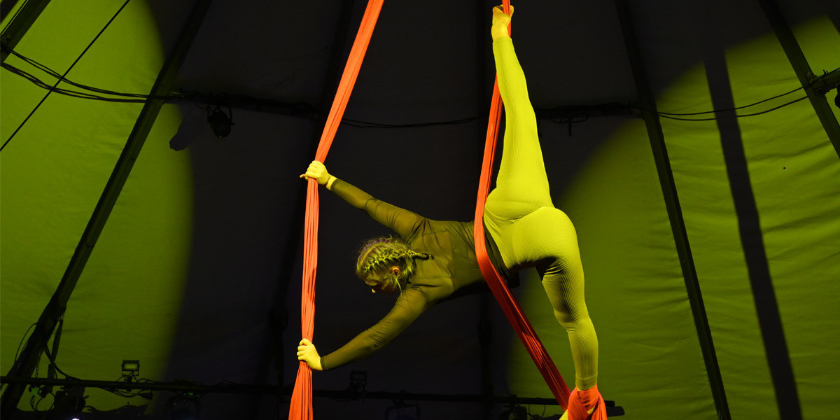 An aerialist performs a vertical split in the air with a red silk.