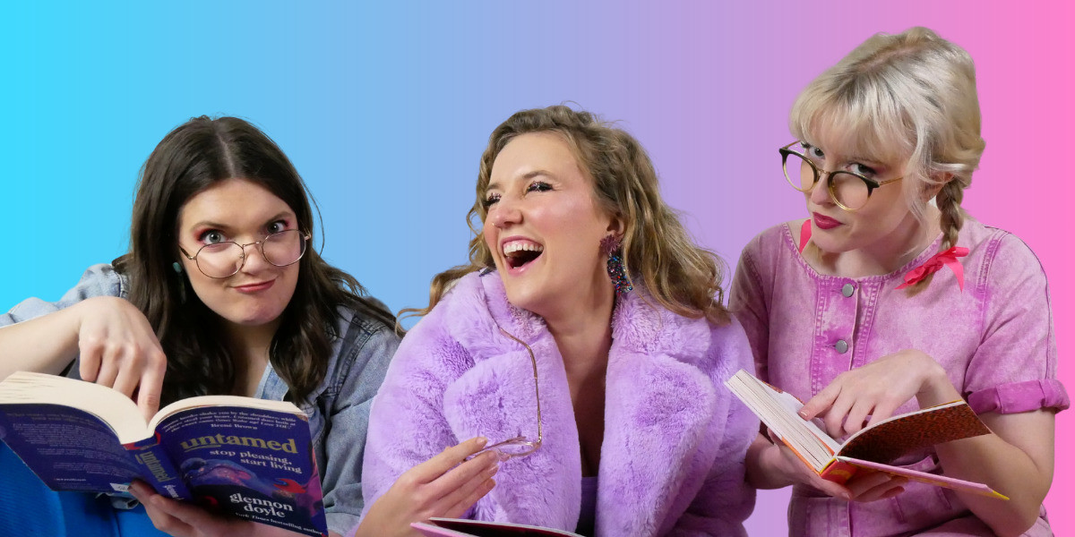 Three white women stand side-by-side with colourful books open in their hands. The woman on the left has dark hair and is wearing a blue denim jacket and glasses, pointing to her dark purple-covered book and pulling a face. The woman in the middle has dark blonde hair and is wearing a fluffy purple coat, and is laughing broadly with her eyes closed. The woman on the right is wearing a pink boiler-suit and her platinum blonde hair is in plaits, and she is wearing glasses, pointing to her book with a bookish expression. Behind them is a gradient of blue, purple and pink.