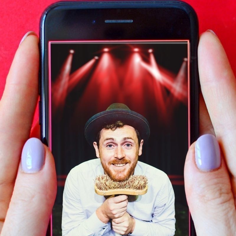 Red background of a smart phone being held by a womans hand with purple nail polish. On the screen of the phone is a mans face with a moustache, beard, brown hair and a pork pie hat. The man has a smile on his face. The mans face is resting on a straw broom.