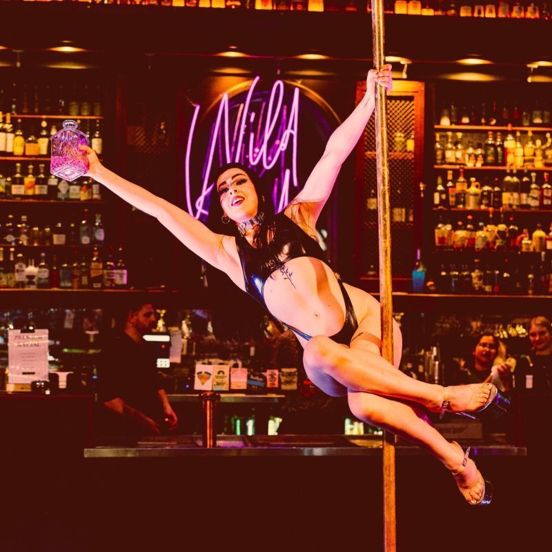 Cleo Rapture wears a shiny black cossie and spins on a pole holding out a bottle of pink liquid on a background of whiskey bottles on shelves.