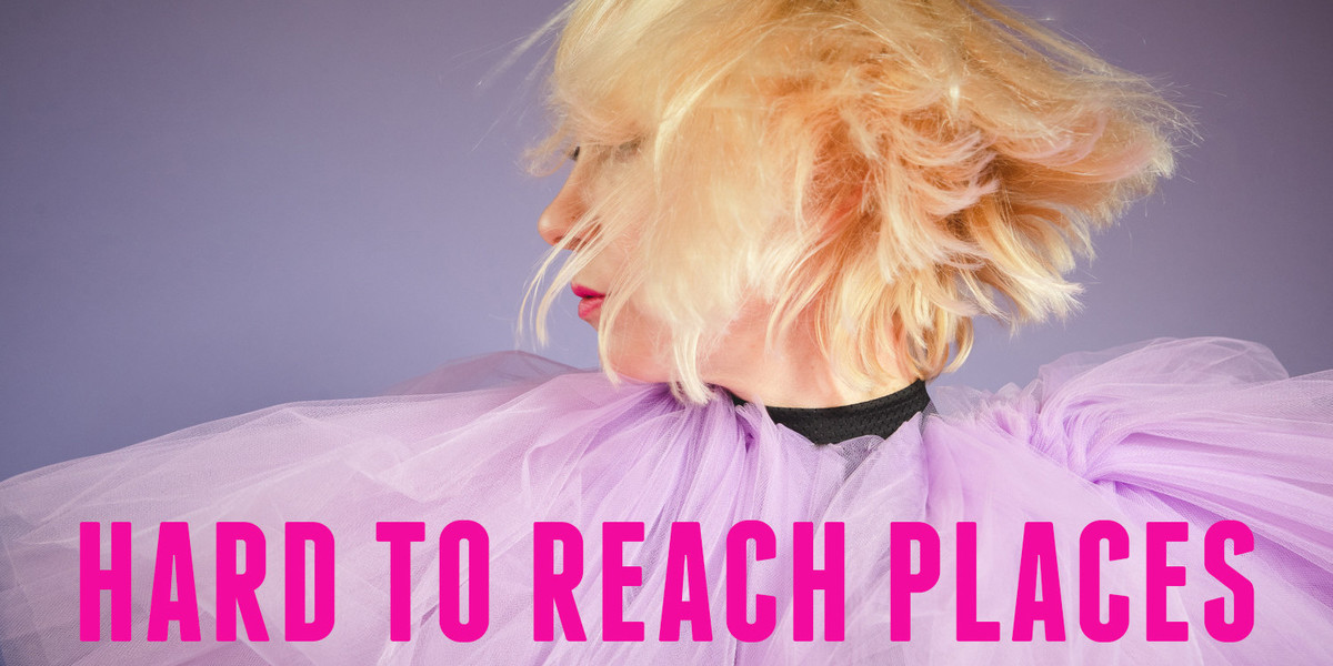 A women is shaking her head, her blonde hair is shown in movement, she is wearing a purple tulle collar. There is pink text at the bottom which read "Hard to Reach Places."