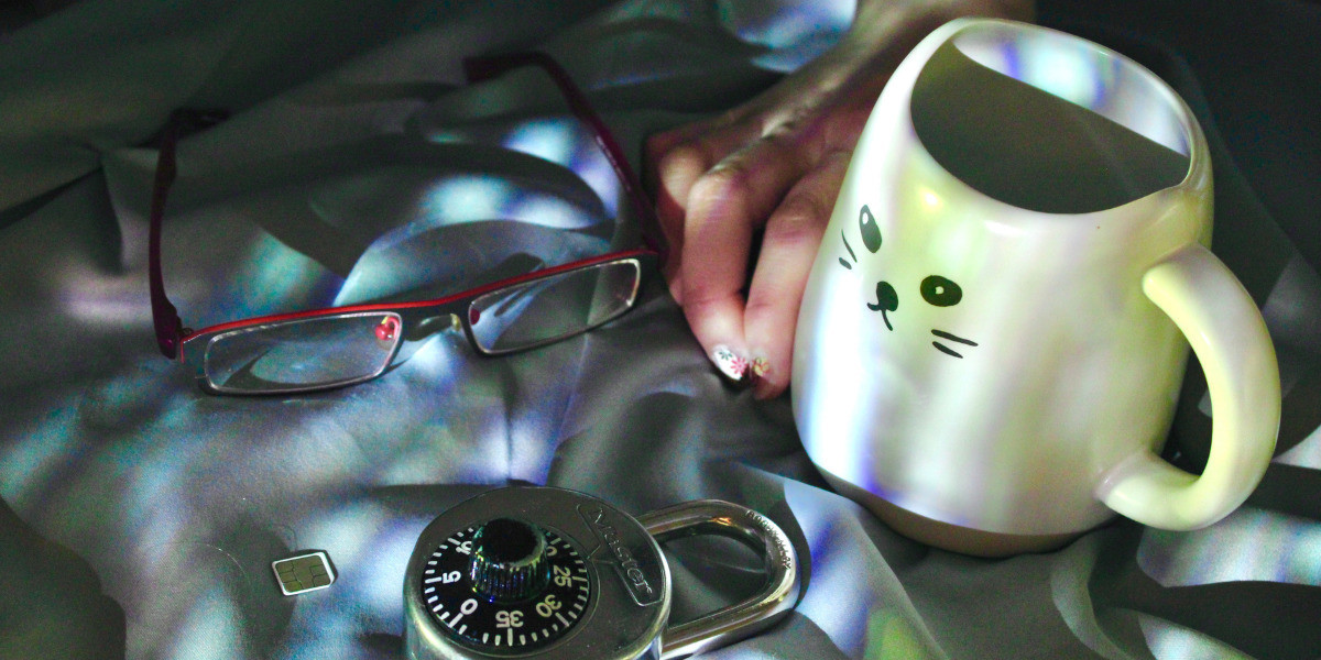 Four objects can be seen laying on a piece of grey fabric. From left to right, they are a pair of glasses, sim card, a coded lock, and a mug. The glasses have dark grey frames and red handles. The sim card is tiny in the shape of a square with a reflective surface. The coded lock is silver and reflective, and has the words "Master" on the front and a black coded spinner with white numbering. The mug is white and has the face of a cat drawn on the front, with two pointed edges on the top to signify ears. A tan-skinned hand with floral nail art is situated in between the glasses and the mug, in a curled position. Tinges of blue, purple and green are seen reflected onto the objects.