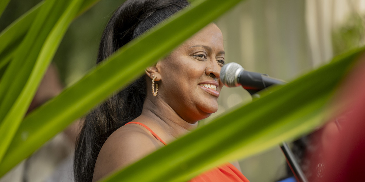 A Women, person of colour, wearing orange dress,smiling in front of a microphone sorounded by plants