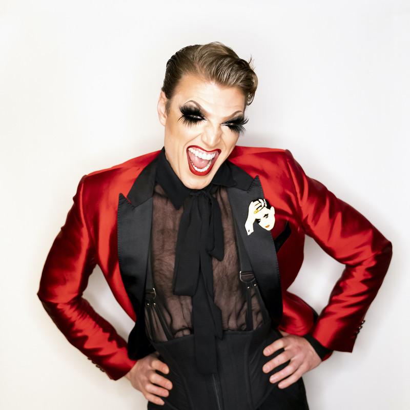 Man stands with hips on his hands in a Red shiny suit jacket, Black sheer shirt, waist cincher, and large brooch of himself on the jacket lapel. He has a full face of makeup on, with blonde quiff and bright red lipstick. His mouth is open like he is smiling and screaming excitedly baring his perfectly straight white teeth.