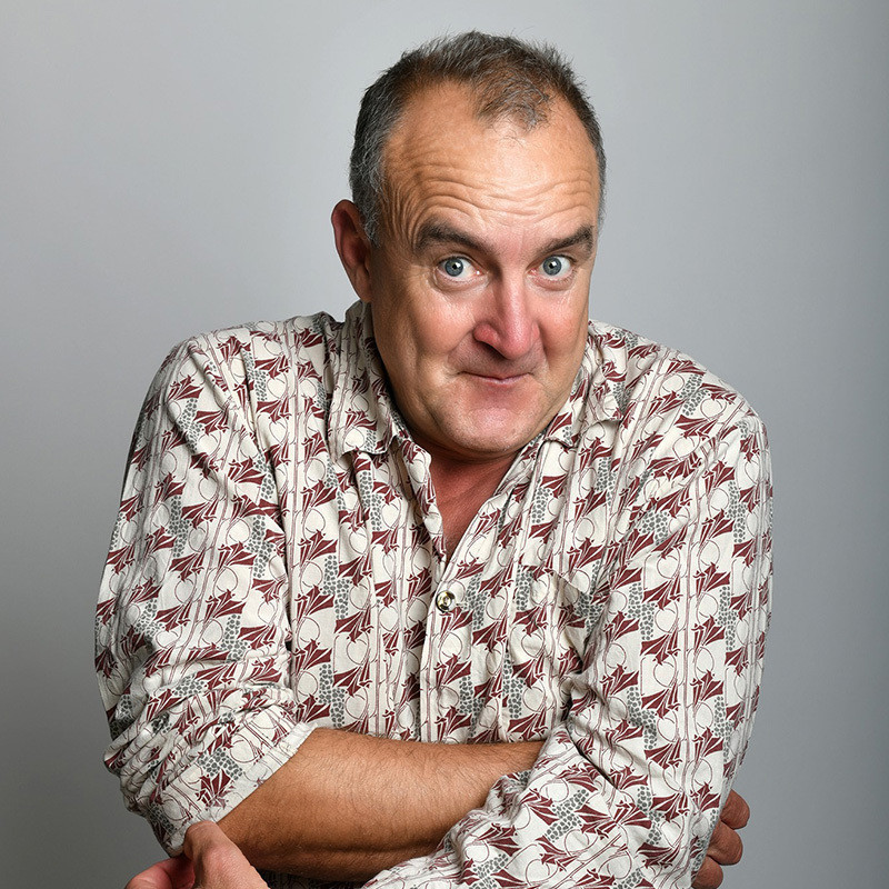 A man with greying hair looks at the camera with a cheeky raised eyebrow. He wears a white shirt with small red and blue patterns, the background is grey.