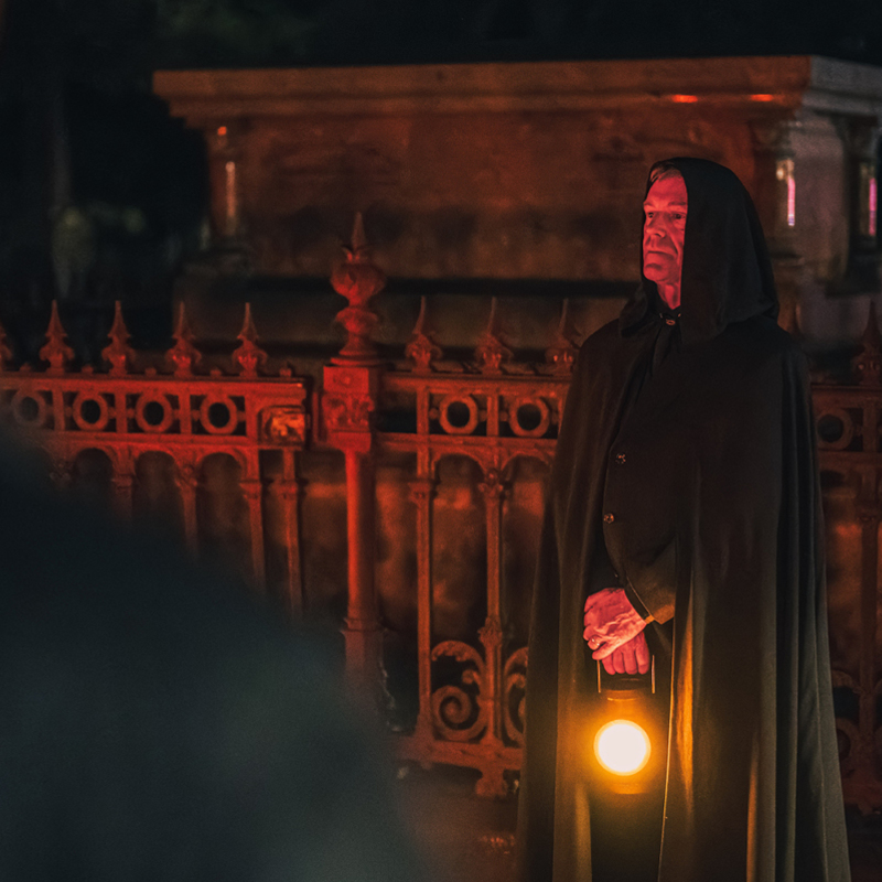Cloaked Man stands in front of grave holding lantern. In the foreground is the blurred silhouette of tour guests.