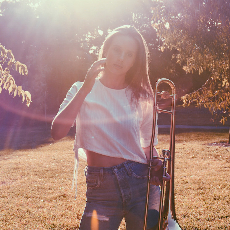 A female with long brown hair, wearing a white shirt and blue jeans, standing outside amongst the trees and grass, with the afternoon sun shining through. They are holding a trombone in their left hand, their right arm is raised.