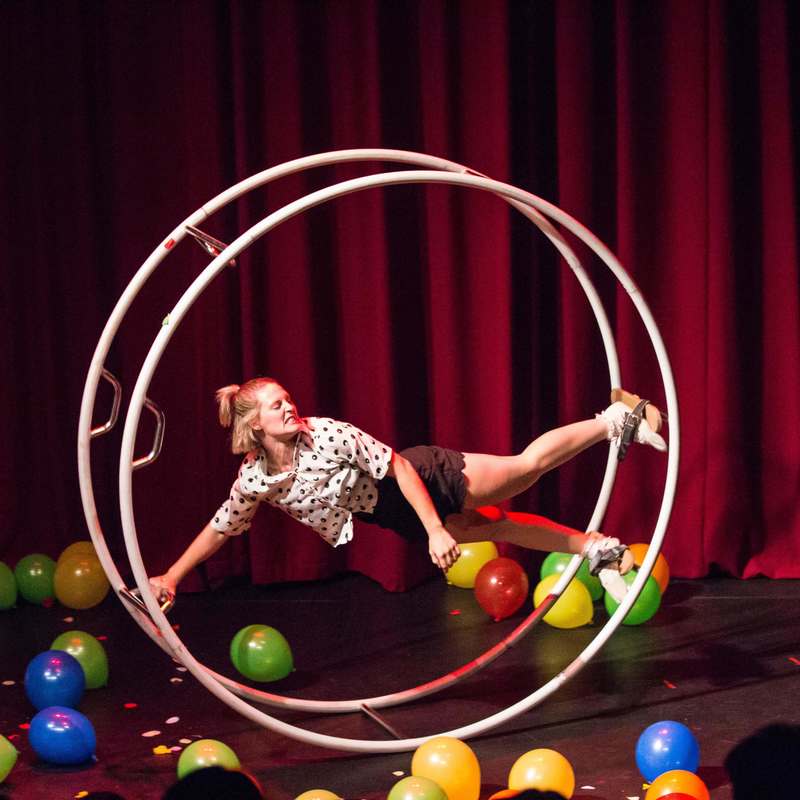 One person is held horizontal strapped into a giant white wheel. The ground is covered in balloons and there is a red curtain in the background.