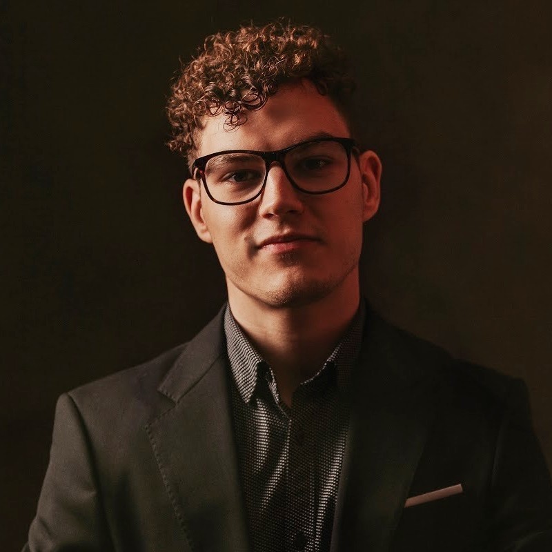 Kelsey De Almeida: Unlikely - A headshot of a man with short curly brown hair and dark rimmed glasses. He wears a suit jacket and a shirt, the background is a dark grey.