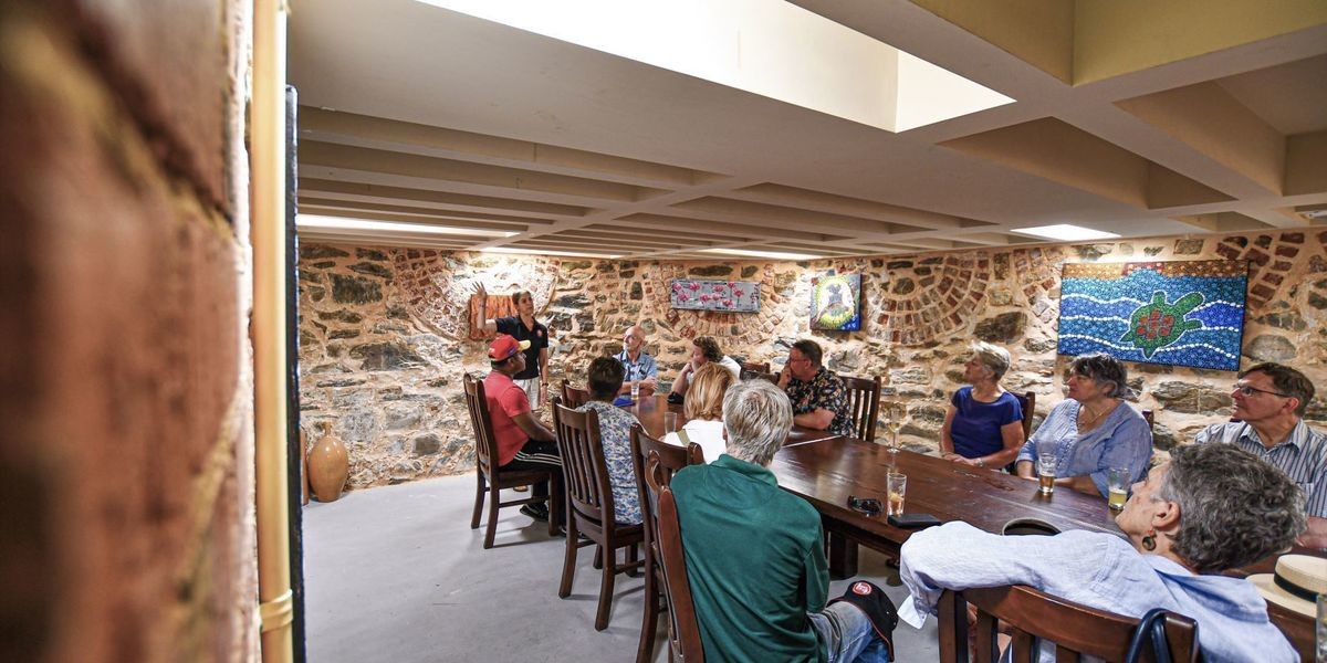 A tour guide talks to their group who are seated around a table, in a cellar.