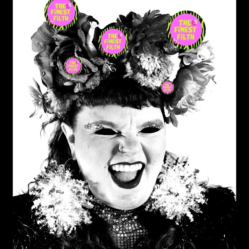 The Finest Filth Variety Hour - The image is a black and white close up photo of Annie Schofield smiling. Their eyes have been coloured in black to create a 'freaky' effect. Annie wears huge tinsel earrings, a headpiece with pom poms, baubles, butterflies and a little doll and barbie arm. They are wearing a high collared sparkly, gold jumpsuit and a pink and purple tinsel jacket.