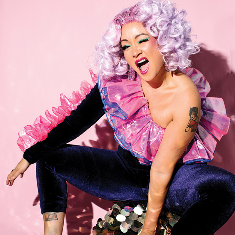 Smashed - The Brunch Party - A portrait shot featuring Victoria Falconer, the host of Smashed, who is a mixed-race cis-woman, sitting on a gold sequin-covered seat, set against a light pink background. She is wearing an outrageously excellent one-shouldered catsuit made of purple velvet, with blue and pink iridescent tulle frills along one arm, and around the neckline. She has a tattoo of a pin-up in boxing gloves on her exposed shoulder, and is also sporting a glittery, curly lavender-purple wig, bright pink lipstick, with an open-mouthed smile and winking cheekily at the camera.